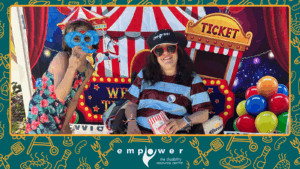 Empower N L Board Member Mandy Penney poses in front of the carnival wall, with a masked friend. The Empower N L logo is centered at the bottom.
