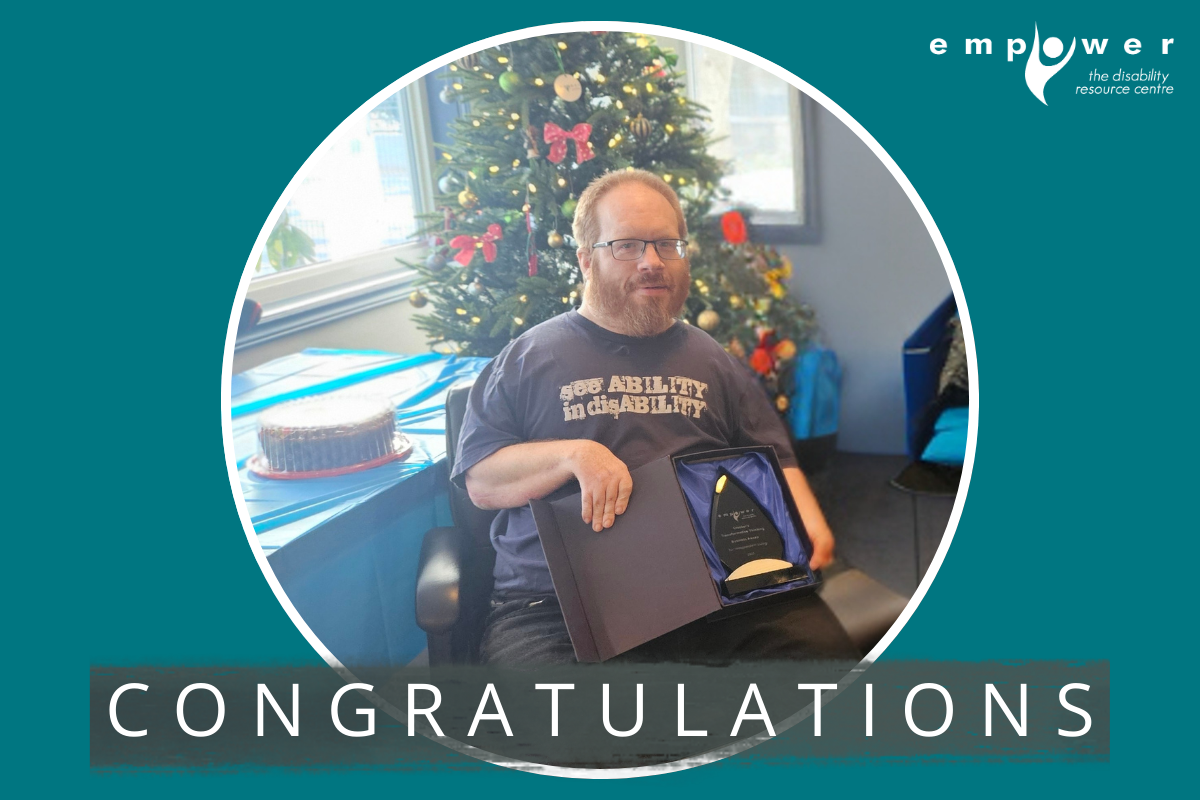 Sheldon Crocker holds his Business Award for Independent Living while sitting in front of a Christmas tree. Text reads Congratulations! The Empower N L logo is in the upper right corner.